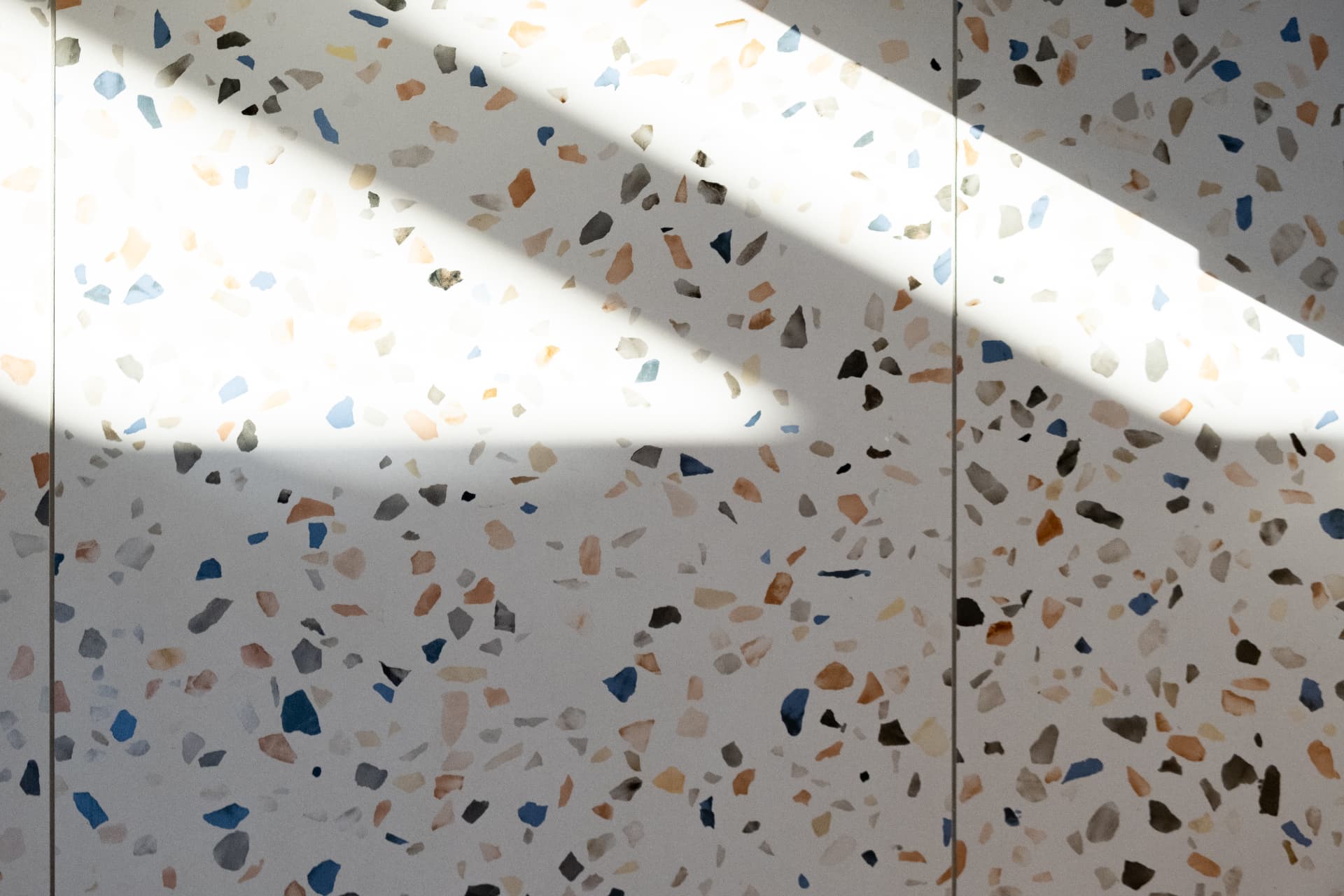 Daylight bands falling across a pale terrazzo surface with blue and ochre aggregate