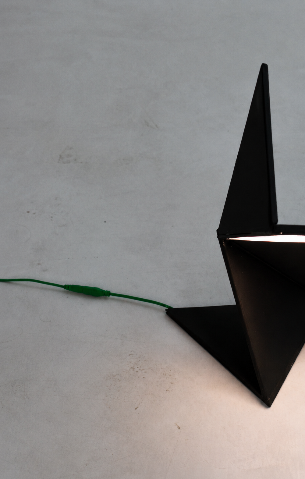 Photograph of the folded black lamp glowing on a concrete surface with a green cable
